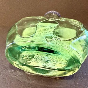Puede incluir: Una botella de vidrio transparente verde con una tapa plateada. La botella tiene una forma redondeada con una superficie texturizada y contiene un l&iacute;quido verde. El texto es visible en la superficie de la botella.