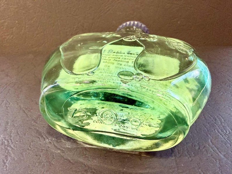 Puede incluir: Una botella de vidrio transparente verde con una tapa plateada. La botella tiene una forma redondeada con una superficie texturizada y contiene un l&iacute;quido verde. El texto es visible en la superficie de la botella.