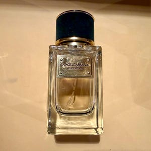 Puede incluir: Un frasco de perfume rectangular de vidrio con una tapa de terciopelo verde oscuro y una banda dorada. El frasco tiene una etiqueta dorada con las palabras "Dolce & Gabbana Velvet Vetiver". El perfume en su interior es de color &aacute;mbar claro.