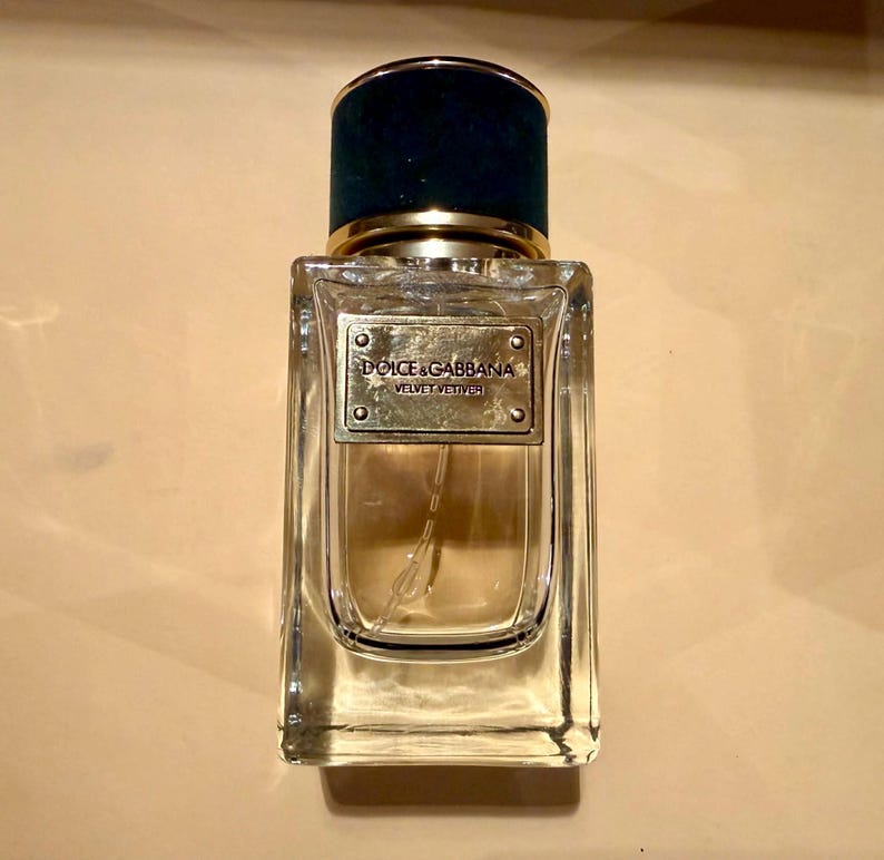 Puede incluir: Un frasco de perfume rectangular de vidrio con una tapa de terciopelo verde oscuro y una banda dorada. El frasco tiene una etiqueta dorada con las palabras "Dolce & Gabbana Velvet Vetiver". El perfume en su interior es de color &aacute;mbar claro.
