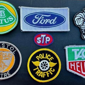 May include: A collection of nine embroidered patches featuring various logos and designs. The patches include logos for Lotus, Ford, STP, Bristol, Police Traffic, TAG Heuer, and a Michelin Man.