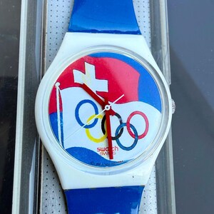 May include: A Swatch wristwatch featuring a white case and a blue strap. The watch face displays a graphic of the Olympic rings and a Swiss flag design with red, white, and blue colors. The watch has red hour, minute, and second hands.