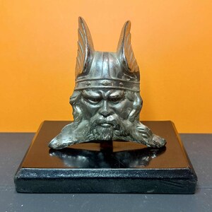 Può includere: Una scultura a busto vichingo color argento con un volto dettagliato e un elmo alato, montata su una base rettangolare nera. La scultura ha un aspetto invecchiato, che suggerisce un pezzo antico o vintage. Lo sfondo è arancione.