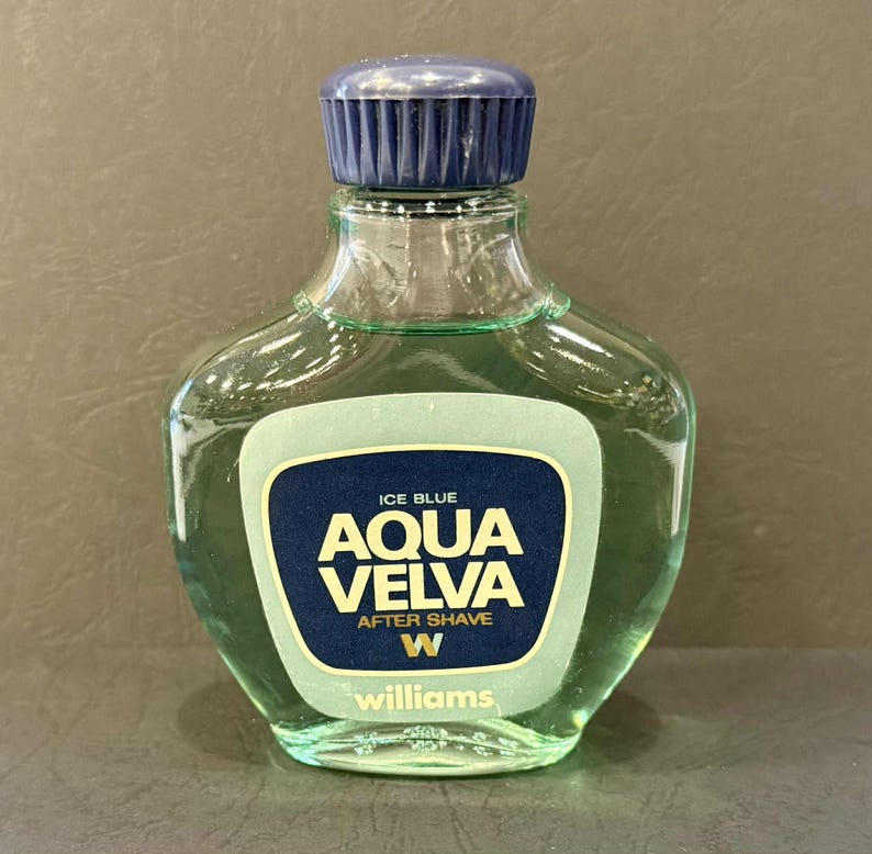 Puede incluir: Botella de cristal transparente de loci&oacute;n para despu&eacute;s del afeitado Aqua Velva. La botella tiene un tap&oacute;n azul oscuro y una etiqueta con las palabras "Aqua Velva" en letras doradas sobre un fondo azul oscuro. Contiene un l&iacute;quido verde claro.