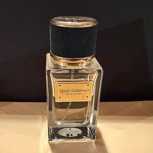 Puede incluir: Un frasco de perfume de vidrio transparente con un tap&oacute;n de terciopelo negro y detalles dorados. El frasco tiene forma rectangular y una etiqueta dorada con la inscripci&oacute;n "DOLCE & GABBANA VELVET VETIVER".