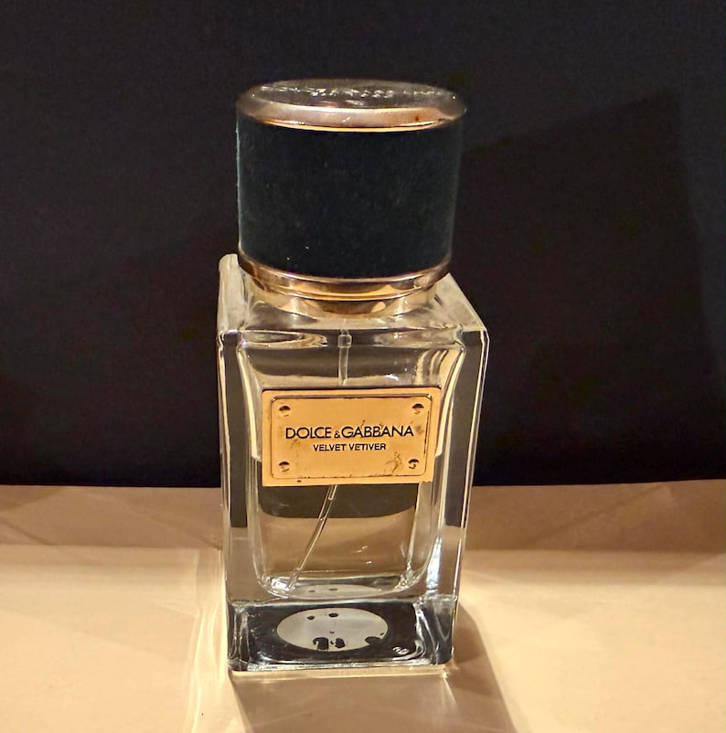 Puede incluir: Un frasco de perfume de vidrio transparente con un tap&oacute;n de terciopelo negro y detalles dorados. El frasco tiene forma rectangular y una etiqueta dorada con la inscripci&oacute;n "DOLCE & GABBANA VELVET VETIVER".