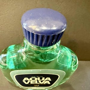 Puede incluir: Una botella de vidrio verde de loci&oacute;n para despu&eacute;s del afeitado Aqua Velva con una tapa azul oscuro. La botella tiene una forma distintiva y una etiqueta con el nombre de la marca. El l&iacute;quido del interior parece ser de color verde claro.