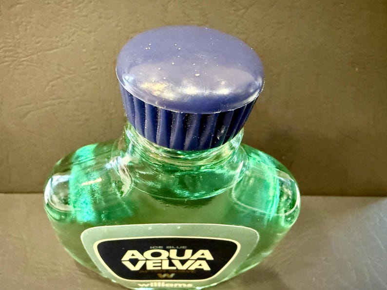Puede incluir: Una botella de vidrio verde de loci&oacute;n para despu&eacute;s del afeitado Aqua Velva con una tapa azul oscuro. La botella tiene una forma distintiva y una etiqueta con el nombre de la marca. El l&iacute;quido del interior parece ser de color verde claro.