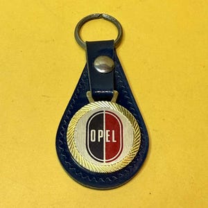 Peut inclure: Un porte-clés Opel vintage avec un support en cuir bleu marine. Le porte-clés présente un emblème en métal doré avec le logo Opel en noir et rouge, et le mot "OPEL" en blanc.