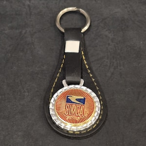May include: A black leather keychain with a silver key ring. The keychain features a round emblem with the word "SIMCA" in gold lettering, a blue and white logo, and a red background.