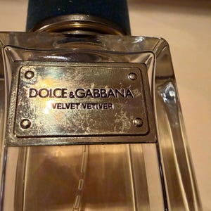 Puede incluir: Un frasco de perfume de vidrio transparente con una placa met&aacute;lica de color plateado. La placa est&aacute; grabada con las palabras "DOLCE & GABBANA VELVET VETIVER". El frasco tiene un tap&oacute;n azul oscuro.