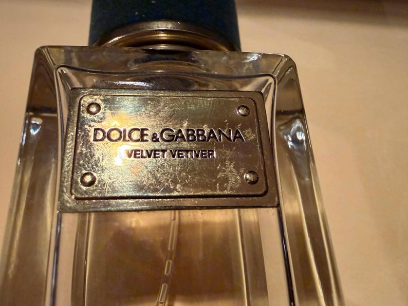 Puede incluir: Un frasco de perfume de vidrio transparente con una placa met&aacute;lica de color plateado. La placa est&aacute; grabada con las palabras "DOLCE & GABBANA VELVET VETIVER". El frasco tiene un tap&oacute;n azul oscuro.