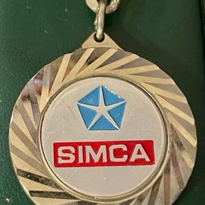 Può includere: Un portachiavi in argento e oro con un cerchio bianco che presenta una stella blu e la parola "SIMCA" in rosso.