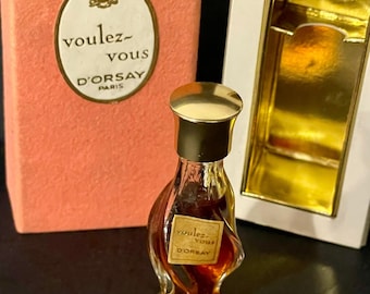 May include: A vintage bottle of "Voulez-vous D'Orsay" perfume in a clear glass bottle with a gold cap. The bottle is shaped like a woman's figure and has a label with the perfume's name on it. The bottle is sitting on a black surface.