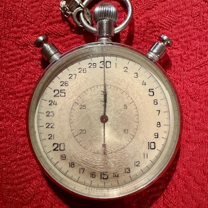 May include: A silver stopwatch with a white face and black hands. The stopwatch has a large outer ring with numbers from 1 to 30, and a smaller inner ring with numbers from 5 to 60. The stopwatch has a chain attached to the top.