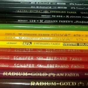 May include: A close-up of a stack of coloured pencils with various labels, including "Extra-Extra" Eberhard Faber, "Johann Faber's "Rafael" N22", "Johann Faber "Diva" 474-N22", "Eberhard Faber Decolor 1827", and "Schiefer Eberhard Faber 22".