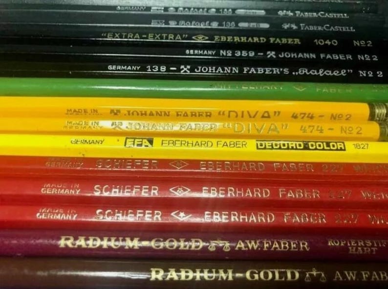 May include: A close-up of a stack of coloured pencils with various labels, including "Extra-Extra" Eberhard Faber, "Johann Faber's "Rafael" N22", "Johann Faber "Diva" 474-N22", "Eberhard Faber Decolor 1827", and "Schiefer Eberhard Faber 22".