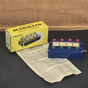 Op de afbeelding: Een vintage Marklin bedieningspaneel model 7072, met een blauwe basis en rode en groene knoppen. Op de gele doos staat "Marklin" en "Control Plate". Er is ook een papier met tekst aanwezig.