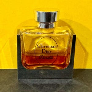 May include: A clear glass bottle of Christian Dior Diorella perfume with a silver cap. The bottle is sitting on a black base.