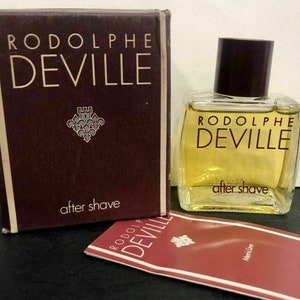 Pode incluir: Uma caixa marrom com o texto "Rodolphe Deville" e "after shave" impresso nela. Um frasco de vidro transparente com uma tampa marrom e o texto "Rodolphe Deville" e "after shave" impresso nele. Um pequeno cartão vermelho e branco com o texto "Rodolphe Deville" impresso nele.