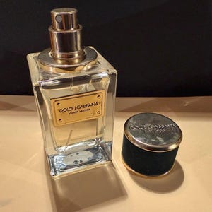 Puede incluir: Un frasco de perfume Dolce & Gabbana Velvet Vetiver de vidrio transparente con tap&oacute;n y etiqueta dorados. El frasco es rectangular con un pulverizador dorado. El tap&oacute;n es negro con borde dorado y el nombre de la marca.