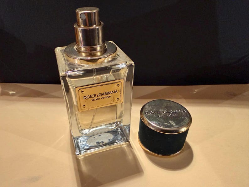 Puede incluir: Un frasco de perfume Dolce & Gabbana Velvet Vetiver de vidrio transparente con tap&oacute;n y etiqueta dorados. El frasco es rectangular con un pulverizador dorado. El tap&oacute;n es negro con borde dorado y el nombre de la marca.