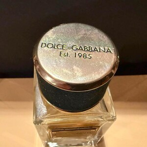 Puede incluir: Un frasco de perfume con una tapa plateada con las palabras "DOLCE & GABBANA Est. 1985". La tapa tiene un borde dorado y se asienta sobre un frasco de vidrio transparente y rectangular. La botella tiene un cuello negro.
