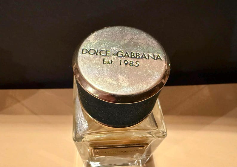Puede incluir: Un frasco de perfume con una tapa plateada con las palabras "DOLCE & GABBANA Est. 1985". La tapa tiene un borde dorado y se asienta sobre un frasco de vidrio transparente y rectangular. La botella tiene un cuello negro.