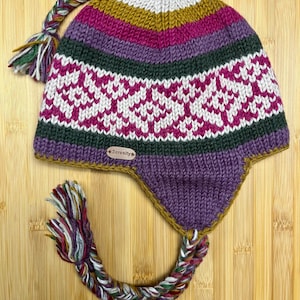 May include: A knit earflap hat featuring horizontal stripes in mustard yellow, magenta, dark green, and white. The hat has a white and magenta geometric pattern, a braided tassel at the top, and two braided tassels at the earflaps. A small tan tag reads Serenity.