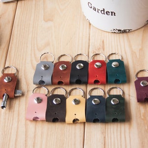 May include: A collection of leather keychains in various colors, including brown, gray, black, red, teal, pink, dark brown, yellow, navy, and dark green. Each keychain features a metal ring and a snap closure. The keychains are arranged on a wooden surface.