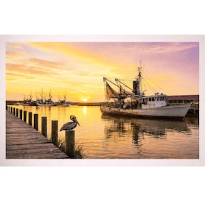 May include: A scenic photograph of a harbor at sunset. A pelican perches on a wooden piling in the foreground. Fishing boats are docked in the water, with a vibrant orange and yellow sky in the background.