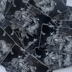 May include: A stack of black and white stickers with a graphic of a snarling mouth with teeth and a web-like design. The stickers are printed on a textured, slightly distressed background.