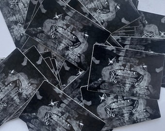 May include: A stack of black and white stickers with a graphic of a snarling mouth with teeth and a web-like design. The stickers are printed on a textured, slightly distressed background.