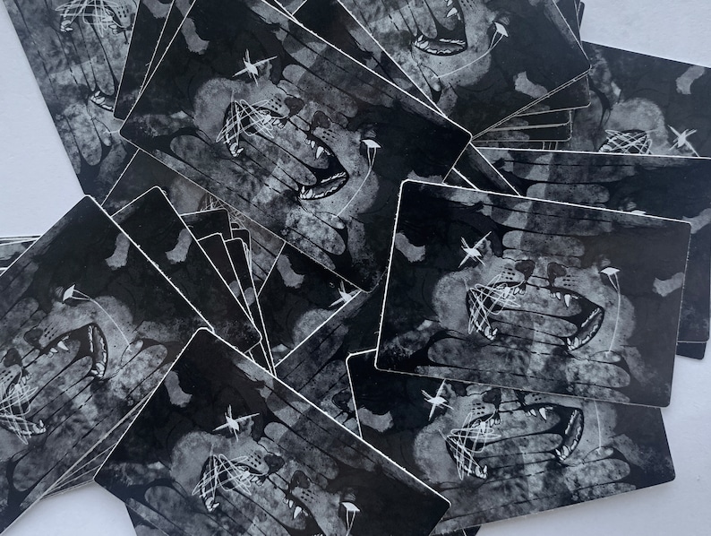 May include: A stack of black and white stickers with a graphic of a snarling mouth with teeth and a web-like design. The stickers are printed on a textured, slightly distressed background.