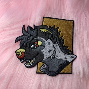 May include: Embroidered patch depicting a stylized hyena head in black, grey, and tan. The hyena has a detailed design with visible teeth and a red nose. The patch is set against a gold rectangular background.