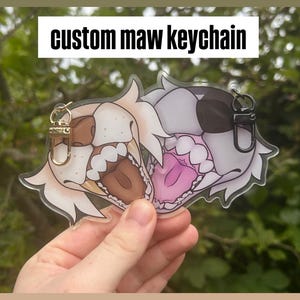 May include: A clear acrylic keychain featuring two cartoon animal faces with open mouths. One is tan and brown, the other is gray and black. The text "custom maw keychain" is above the keychains.