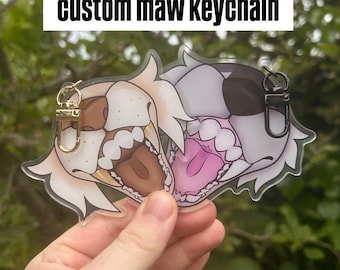 May include: A clear acrylic keychain featuring two cartoon animal faces with open mouths. One is tan and brown, the other is gray and black. The text "custom maw keychain" is above the keychains.