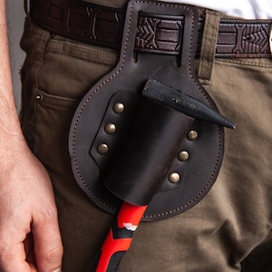 Puede incluir: Un cinturón de herramientas de cuero marrón con un soporte para martillo. El cinturón se lleva alrededor de la cintura de una persona que lleva pantalones caqui.