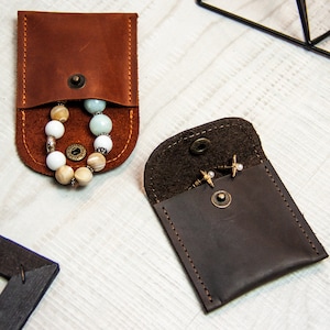 Peut inclure: Deux petites pochettes en cuir avec fermeture à pression. Une pochette est marron et contient un bracelet à perles. L'autre pochette est marron foncé et contient une paire de boucles d'oreilles.