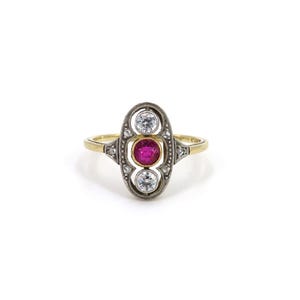 Könnte beinhalten: Ein Ring im Vintage-Stil mit einem goldenen Band und einem oval geformten Mittelstück. Das Mittelstück hat einen zentralen Rubin-Edelstein, der von zwei klaren, rund geschliffenen Steinen flankiert wird. Die Fassung ist mit aufwendigen Metallarbeiten und kleinen Akzentsteinen versehen.