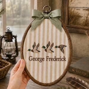 Könnte beinhalten: Ein ovales Stickrahmen-Wandbild mit einem beigeweiß gestreiften Stoffhintergrund. Drei gestickte Stockenten im Flug sind über dem Schriftzug George Frederick in braunem Garn zentriert. Eine salbeigrüne Schleife ist oben am Rahmen befestigt.