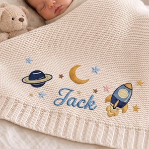 Op de afbeelding: Een crèmekleurige gebreide babydeken met geborduurde ruimtevaartmotieven, waaronder een blauwe planeet met ringen, een gele maansikkel, een blauwe raket en kleine sterren, met de naam Jack geborduurd in blauwe schrijfletters.