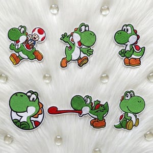 Könnte beinhalten: Sechs gestickte Yoshi-Aufnäher. Jeder Aufnäher zeigt die Figur in verschiedenen Posen, mit grünen, weißen und orangefarbenen Farben. Die Aufnäher stammen aus der Super Mario-Videospielserie.