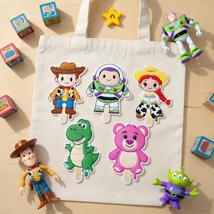 K&ouml;nnte beinhalten: Eine cremefarbene Canvas-Tragetasche mit aufgestickten Charakteraufn&auml;hern von Woody, Buzz Lightyear, Jessie, Rex, Lotso und einem Alien. Spielzeug und Bl&ouml;cke zum Thema Toy Story umgeben die Tasche.