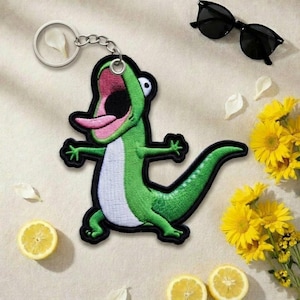 Könnte beinhalten: Ein grüner, gestickter Eidechsen-Schlüsselanhänger mit schwarzer Umrandung, offenem Mund und rosa Zunge. Der Schlüsselanhänger ist an einer silbernen Kette befestigt. Sonnenbrillen, gelbe Blumen und Zitronenscheiben sind ebenfalls im Bild.