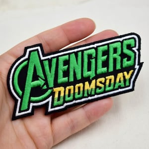 May include: Embroidered patch featuring the text "AVENGERS DOOMSDAY" in green and yellow on a black background. The patch has a white border and is in a rectangular shape. The patch is held in a hand.