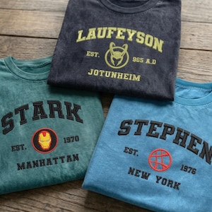 May include: Three crewneck t-shirts in charcoal, teal, and blue. The charcoal shirt features yellow text reading LAUFEYSON EST. 965 A.D. JOTUNHEIM with a helmet icon. The teal shirt has black text STARK EST. 1970 MANHATTAN with an Iron Man mask. The blue shirt has black text STEPHEN EST. 1976 NEW YORK with a circular symbol.