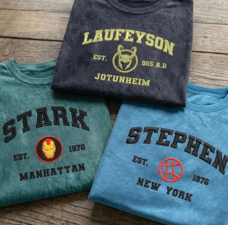 May include: Three crewneck t-shirts in charcoal, teal, and blue. The charcoal shirt features yellow text reading LAUFEYSON EST. 965 A.D. JOTUNHEIM with a helmet icon. The teal shirt has black text STARK EST. 1970 MANHATTAN with an Iron Man mask. The blue shirt has black text STEPHEN EST. 1976 NEW YORK with a circular symbol.