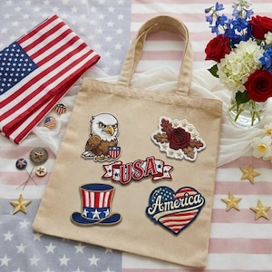 May include: A beige tote bag adorned with patriotic patches, including an eagle, a rose, and a heart-shaped design with the word "America." An American flag and floral arrangement with red roses and blue flowers are also present.
