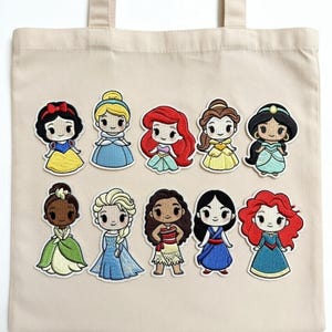 Può includere: Borsa tote beige con toppe ricamate raffiguranti dieci principesse dei cartoni animati in vari abiti colorati. Le principesse includono Biancaneve, Cenerentola, Ariel, Belle, Jasmine, Tiana, Elsa, Vaiana, Mulan e una seconda Ariel.